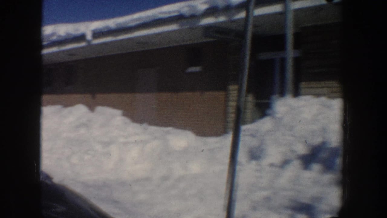 1961 Aspen footage frame showing a long motel-like building with snow on the roof and large snowbanks below a clear blue sky.