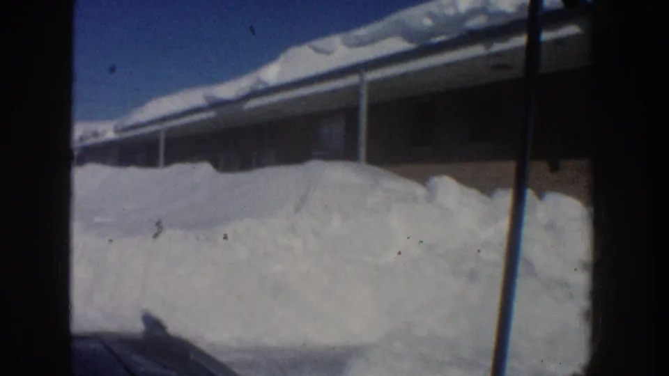 1961 Aspen home-movie frame of a long low lodging building with porch supports, snowbanks, and soft 8mm motion blur.