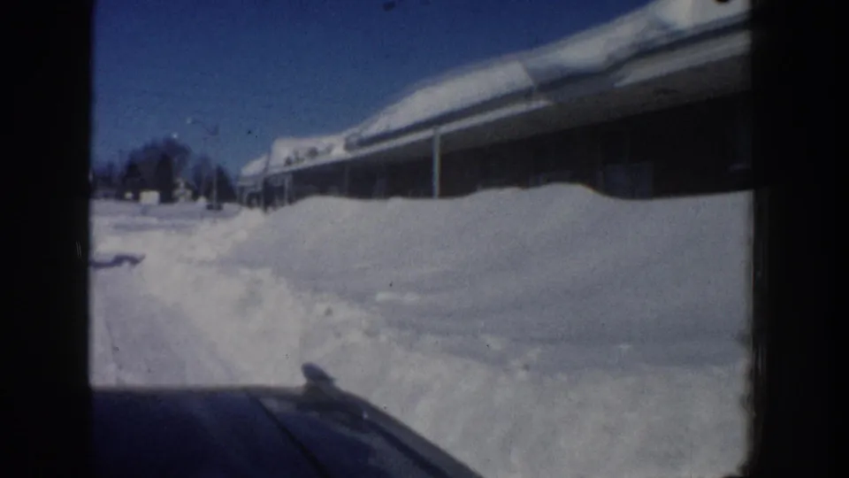 1961 Aspen frame showing a snowy roadside or driveway beside the Holiday Inn building, with roof snow, snowbanks, and distant trees.