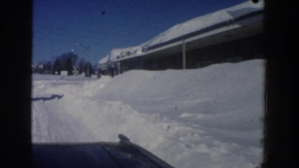 Aspen, Colorado 1961 home-movie frame showing a plowed snowy roadside, a long low building, deep snowbanks, and blue sky.