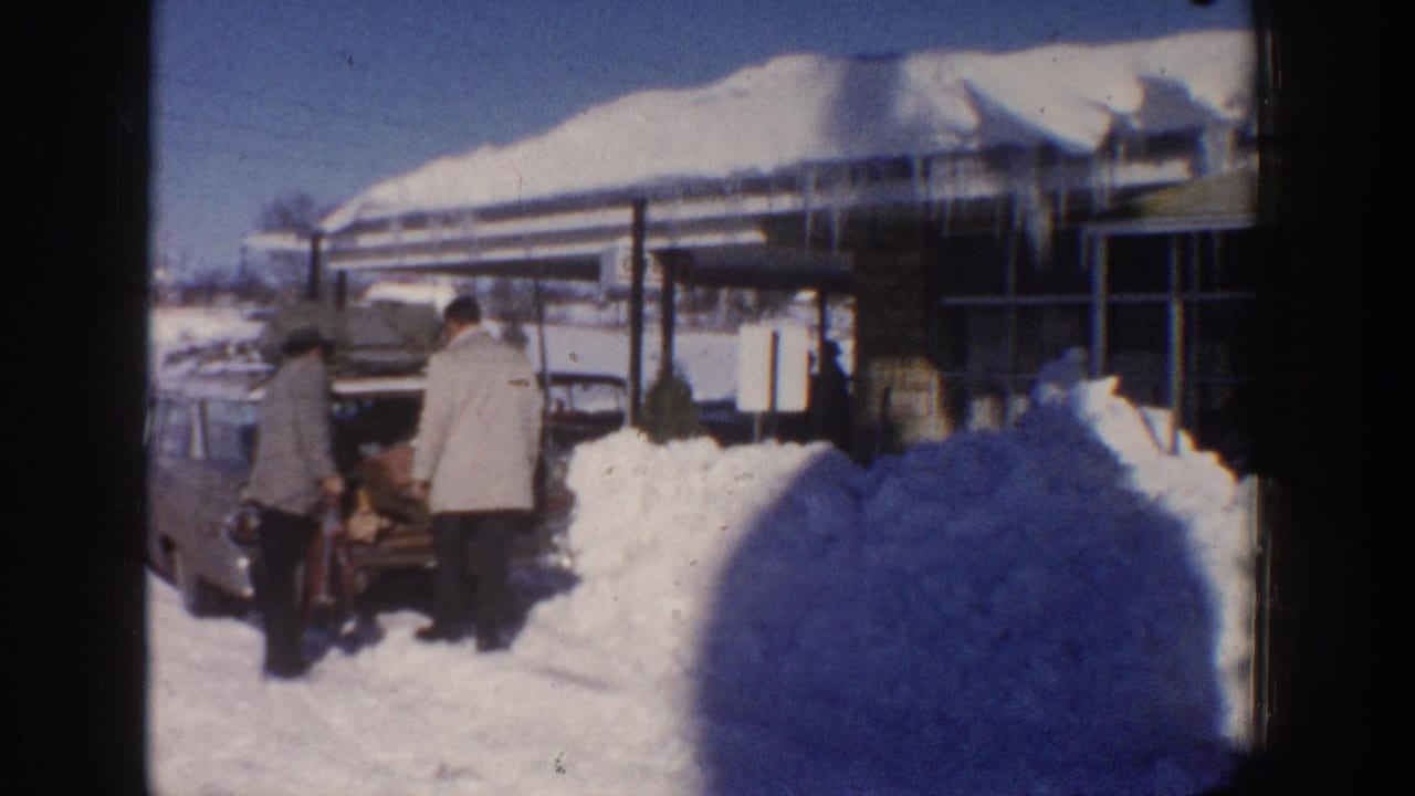 Aspen, Colorado 1961 frame with two adults in winter coats at the rear of a parked car beside deep snow and a snow-laden building.