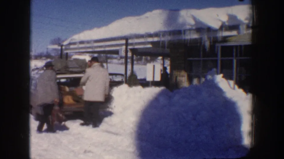 1961 Aspen winter frame showing two adults near an open car trunk, deep roadside snow, and a low building with heavy snow on the roof.