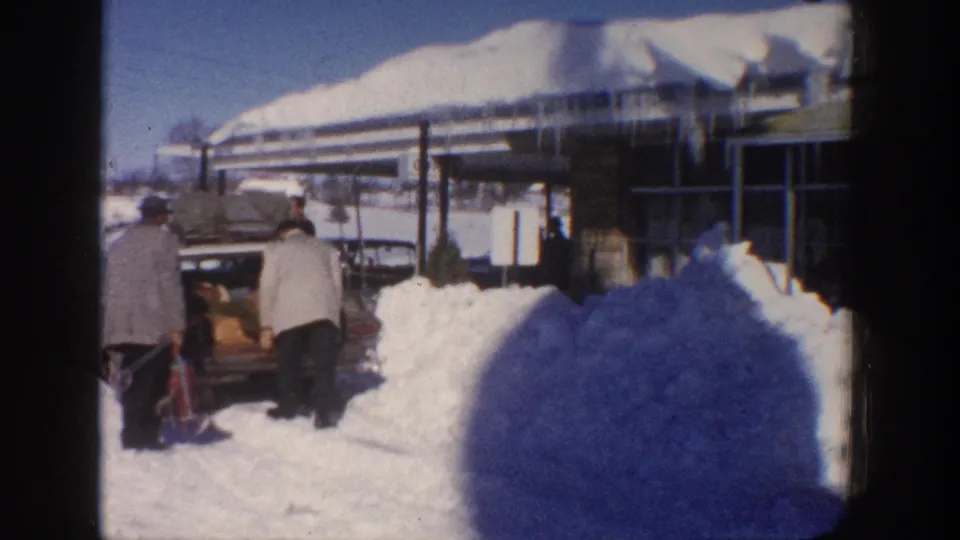 Aspen, Colorado 1961 home-movie frame with two adults by a car, a deep blue-shadowed snowbank, and a building under heavy roof snow.