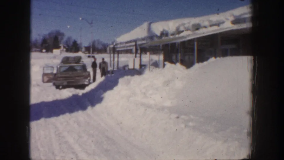 1961 Aspen home-movie frame showing two adults near a station wagon outside a snowed-in building with high snowbanks.