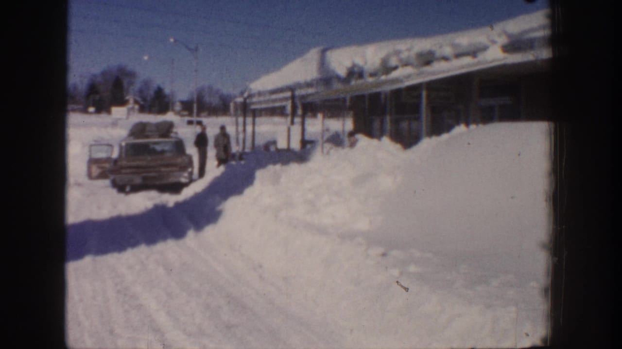 Two adults stand near a station wagon in deep snow beside a low building under bright winter light.