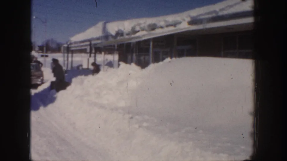 1961 Aspen home-movie frame with a bundled adult near a high snowbank and the station wagon partly visible at the edge.