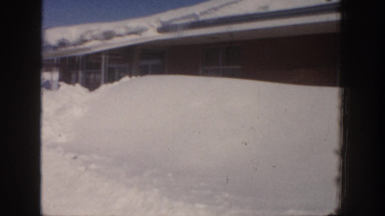 Large snowbank in front of a long low building beneath a blue sky.