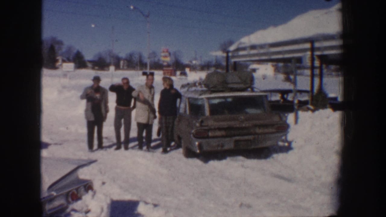Four adults pose or wave near a station wagon parked in a snowy roadside area.