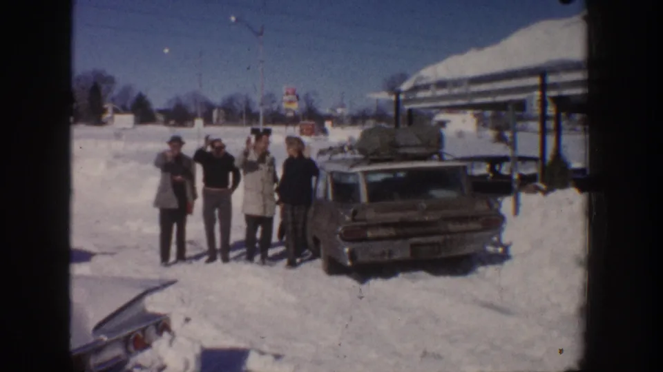 A 1961 Aspen home-movie frame shows adults grouped in snow beside a midcentury station wagon packed for travel.