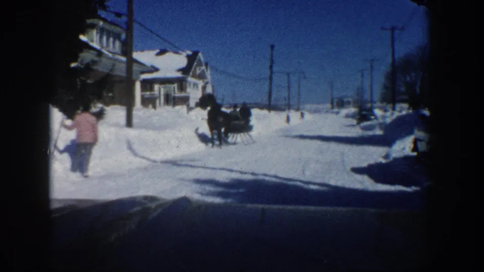 1961 Aspen home-movie frame showing a snowy residential street, a distant horse-drawn sleigh, houses, poles, and a person in pink at left.