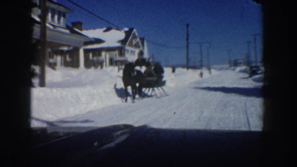 The sleigh travels through a snow-covered Aspen residential street in 1961, framed from inside a vehicle under a clear blue sky.