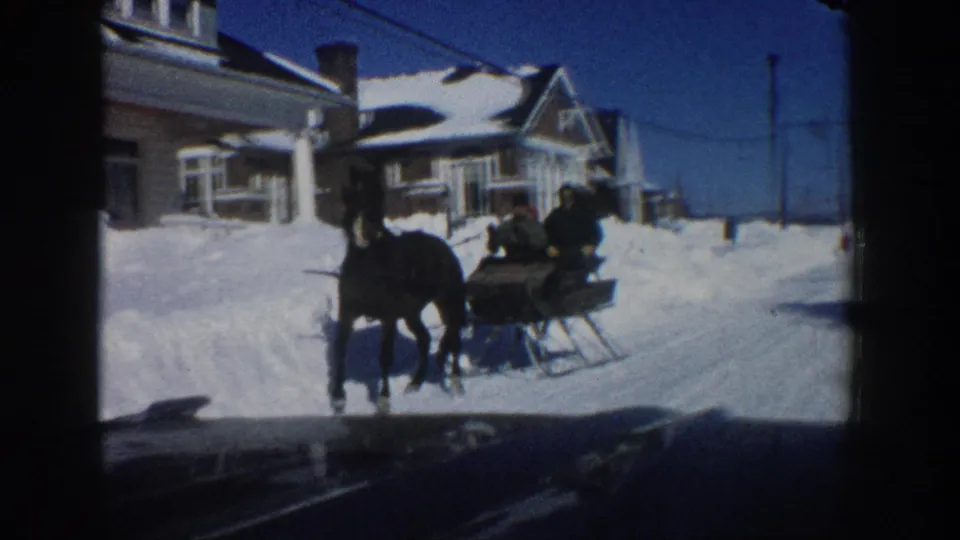 A horse-drawn sleigh with passengers passes closer to the camera on a snowy Aspen street, with houses and deep snowbanks behind it.