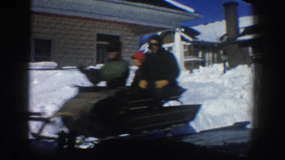 Close 1961 Aspen frame of sleigh passengers passing the camera, with the horse partly visible and snowy houses in the background.
