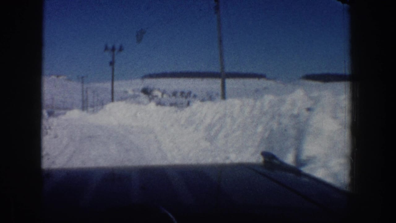 Inside-car view on a plowed winter road in Aspen, Colorado, with deep snowbanks, utility poles, and distant buildings under clear sky.