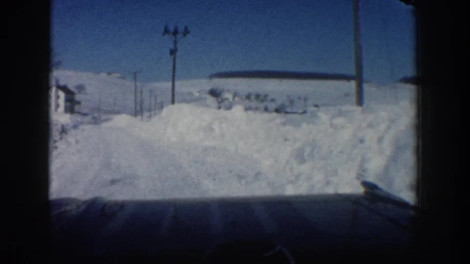 8mm frame from a car traveling a snow-packed Aspen road, showing a high roadside snowbank, utility poles, and a small structure at left.