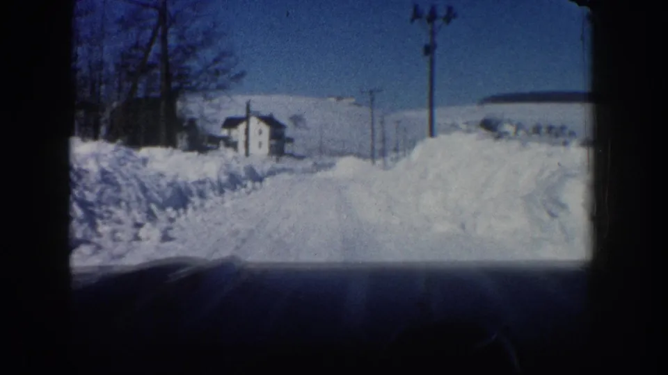 Aspen, Colorado winter driving view from 1961, with a snow-covered road, high banks, utility poles, bare trees, and a small house-like building.