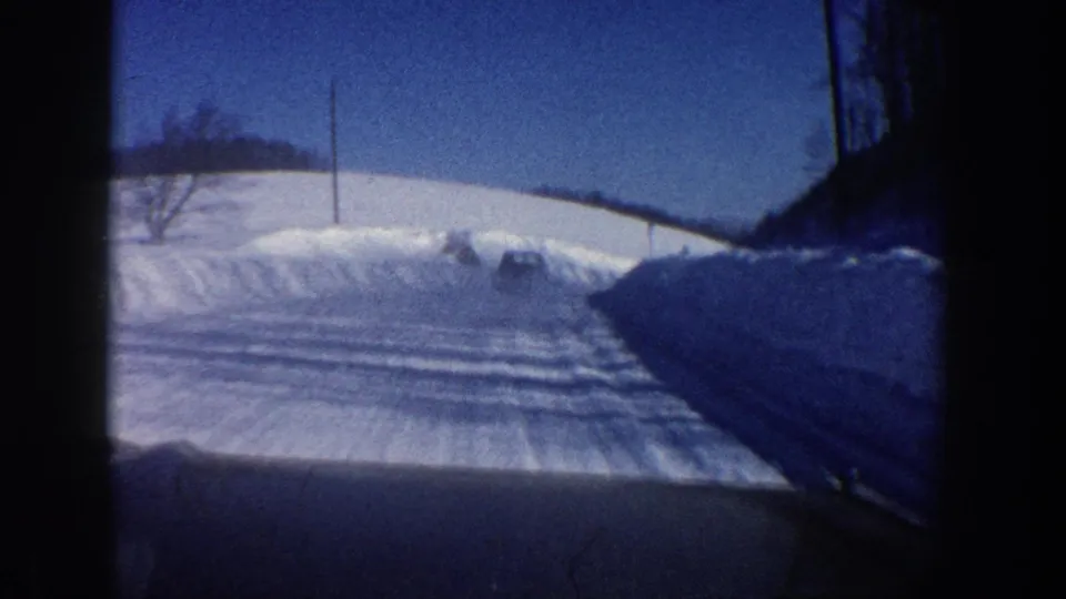 Forward car-window view of a plowed snowy road near Aspen in 1961, with roadside snowbanks and poles.