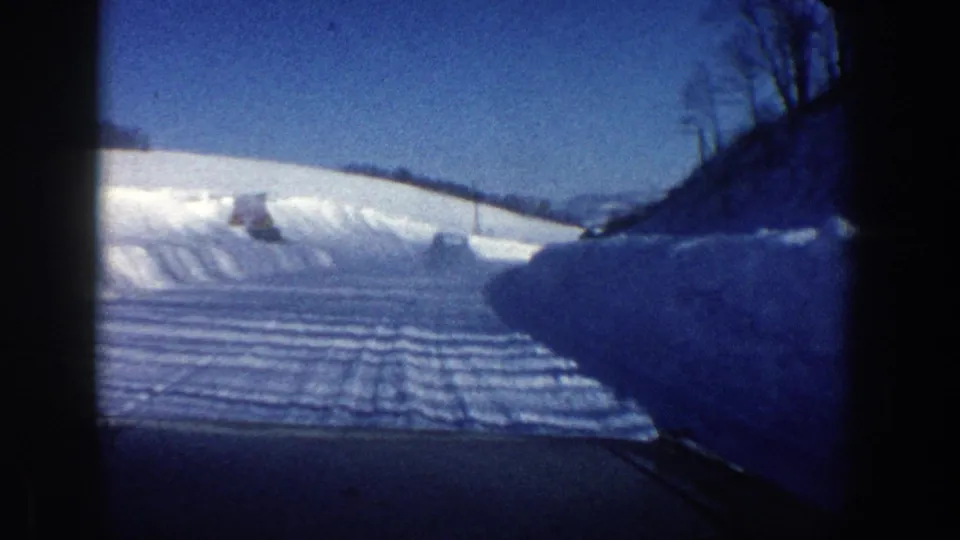 1961 Aspen winter driving frame with a plowed road, high snowbanks, bare trees, and dark windshield borders.
