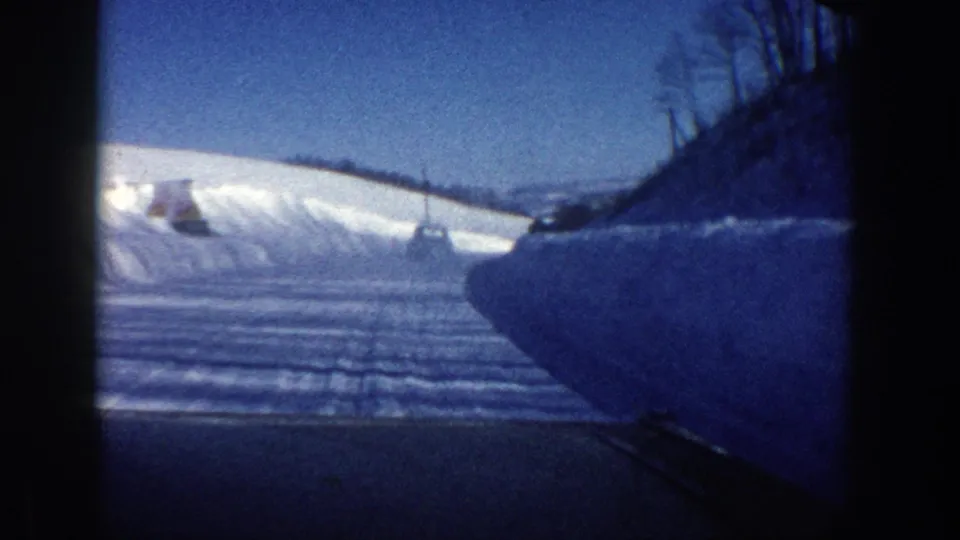 1961 Aspen home-movie view from inside a moving car on a plowed snowy road bordered by high snowbanks and low winter hills.