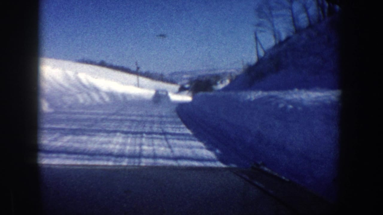Snowy road seen from inside a moving vehicle, with snowbanks and distant low hills.