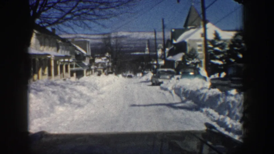 A 1961 Aspen street appears from the moving car, with snowbanks, houses, utility poles, bare trees, evergreens, and parked vintage cars.