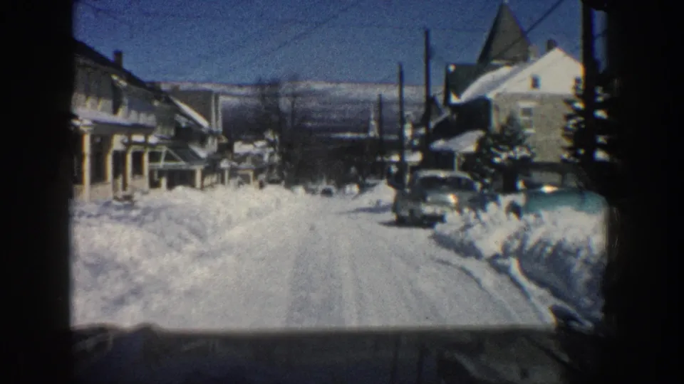 The car proceeds down a snowy Aspen residential street, passing houses, utility poles, snowbanks, and parked mid-century vehicles.