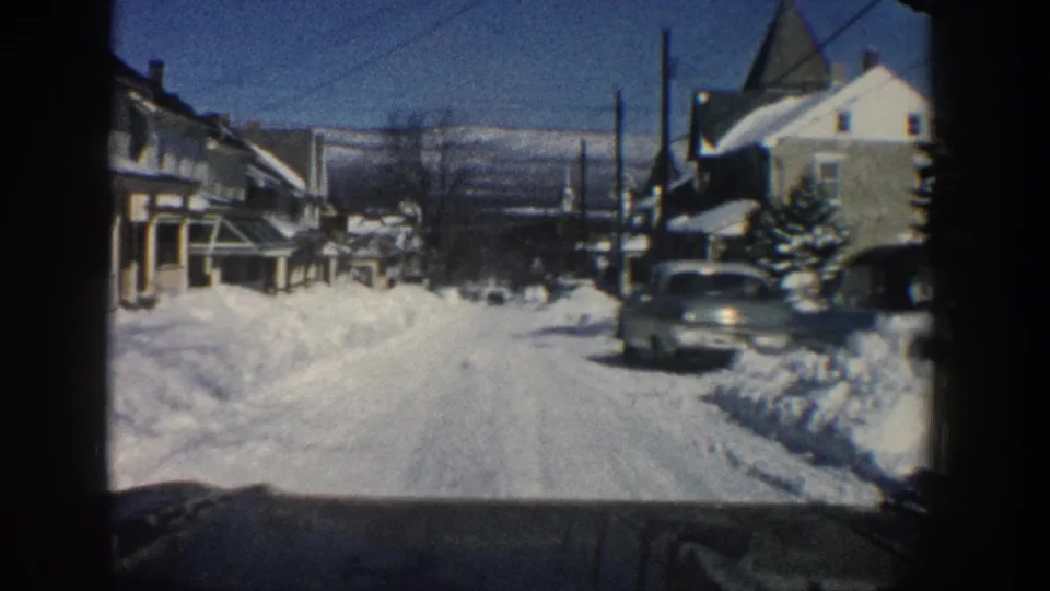 1961 Aspen, Colorado street view from inside a moving car, showing deep snowbanks, houses, parked cars, and distant mountains.