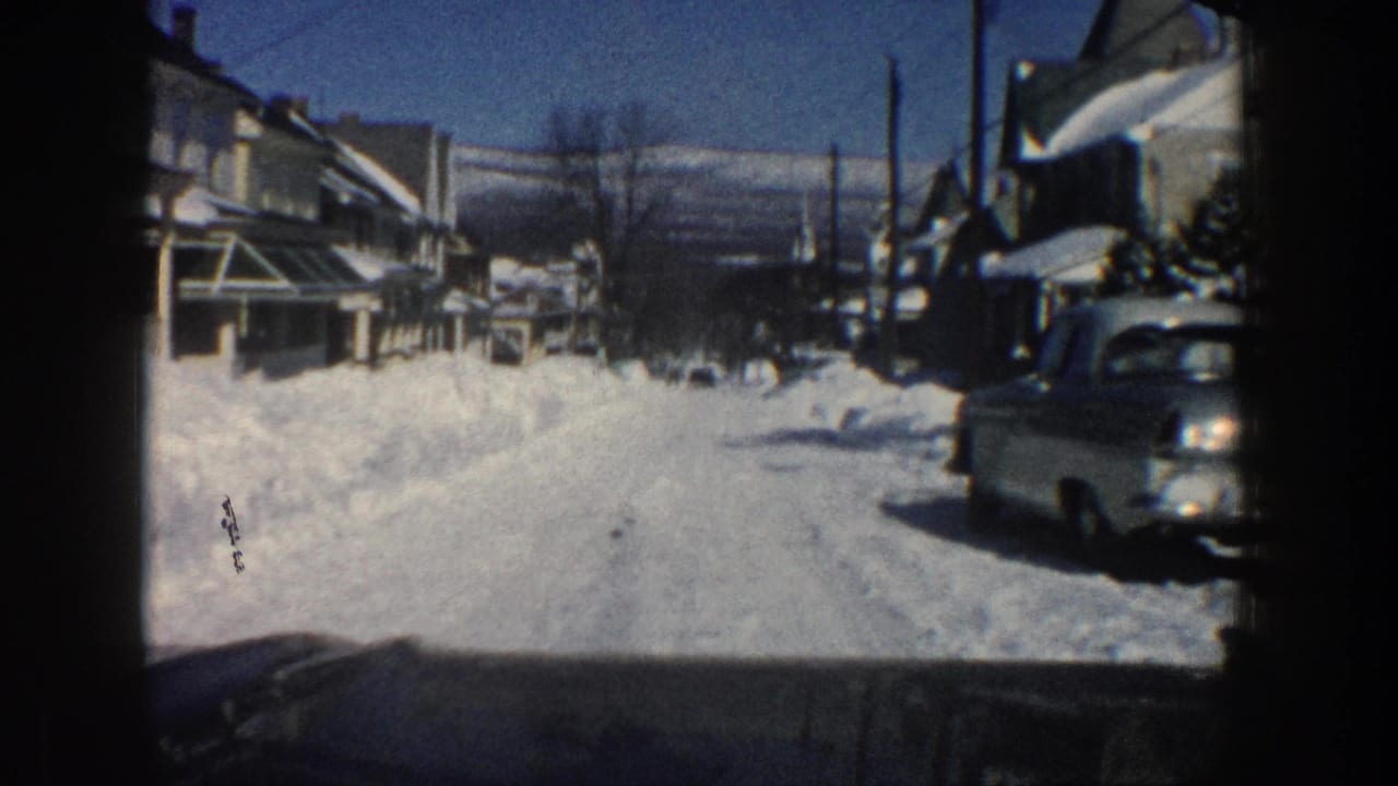 Snowy street with houses, parked cars, utility poles, and a clear winter sky seen from a vehicle.