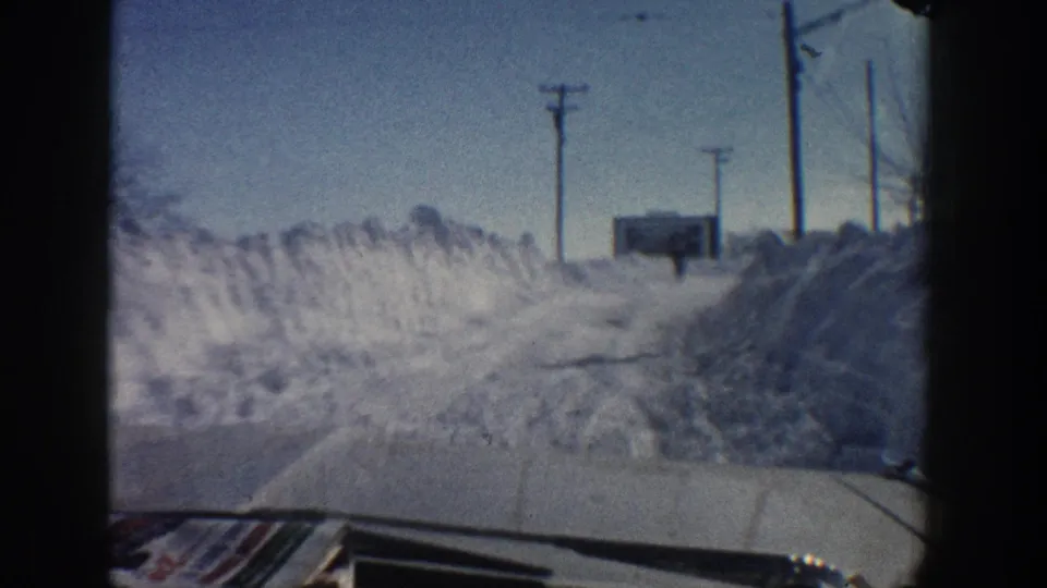 The 1961 Aspen driving shot opens onto a snowbank-lined roadside with utility poles, a distant signboard, blue sky, and the car hood.