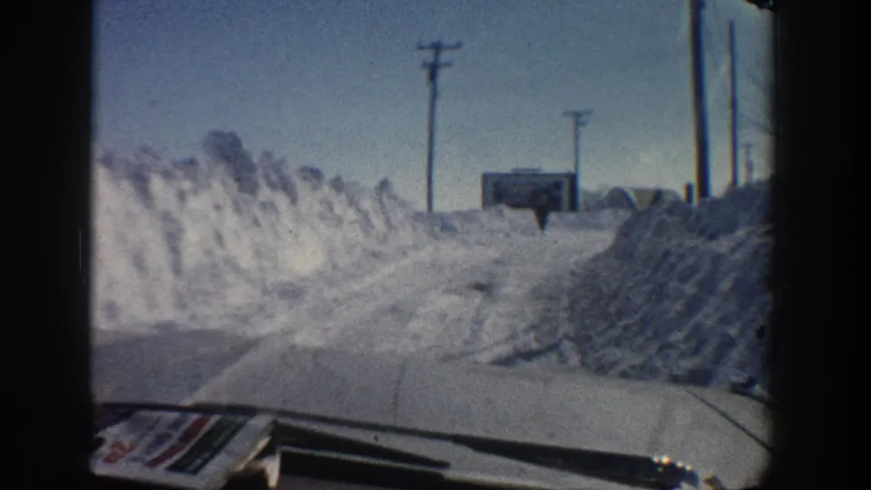 1961 Aspen, Colorado home-movie view from a moving vehicle on a narrow snowy road lined by deep snowbanks and utility poles.