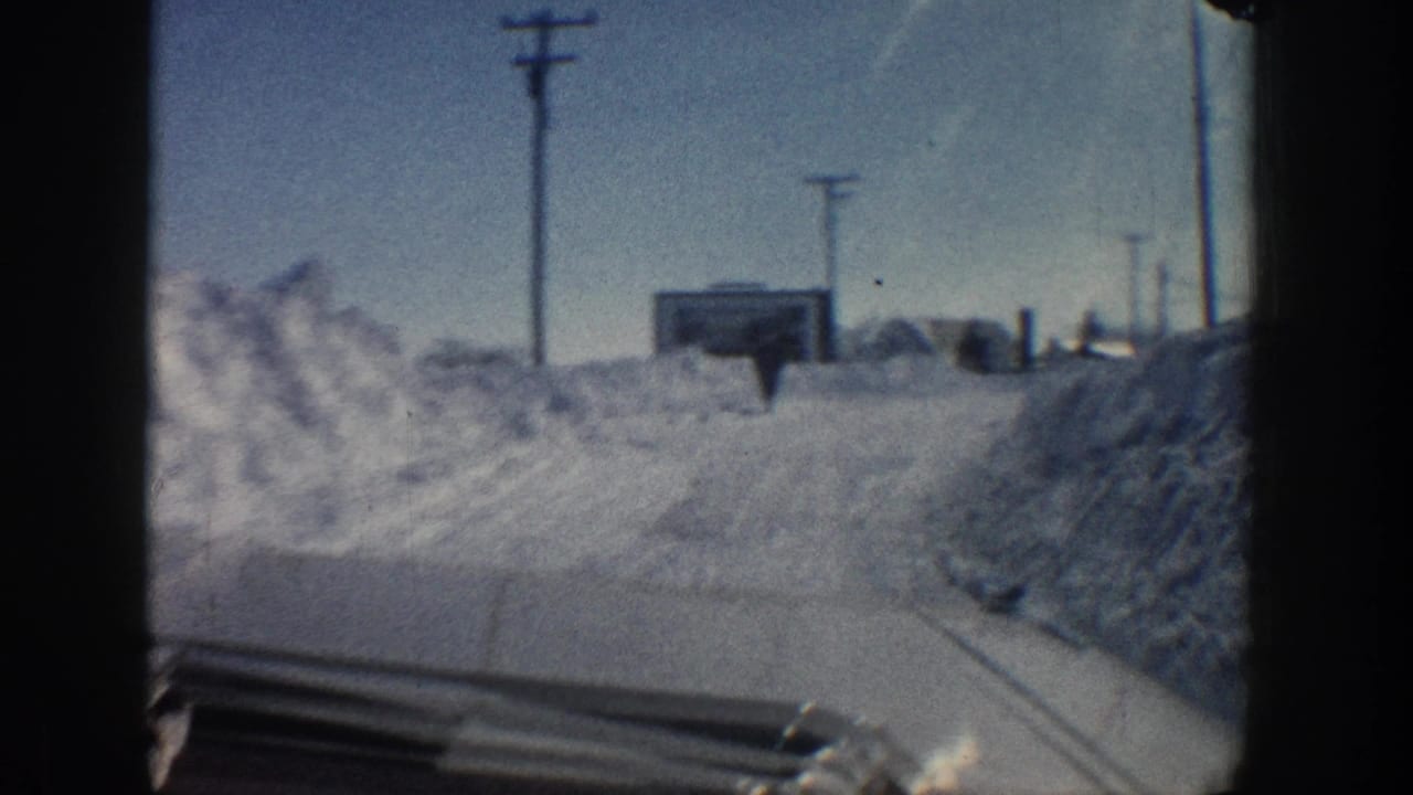 Moving vehicle perspective on a snowy Aspen road in 1961, with deep plowed banks and utility poles receding along the roadside.
