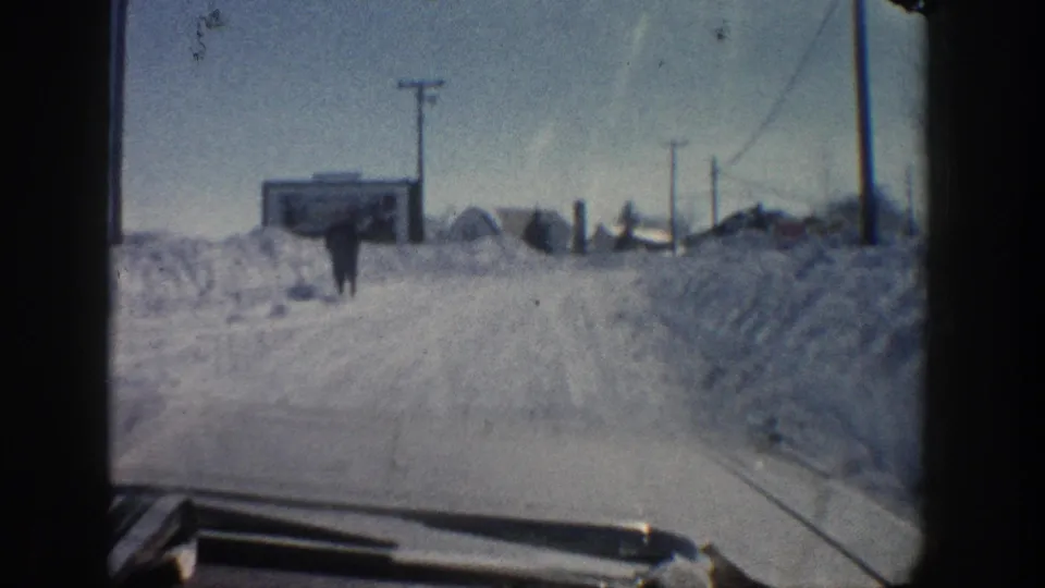 1961 Aspen winter road view from a car, showing a distant pedestrian walking near tall snowbanks and roadside buildings.