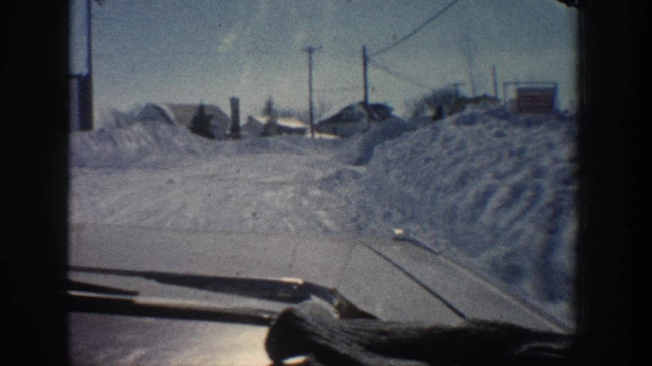 1961 Aspen home-movie view through a car windshield along a snow-covered road bordered by high snowbanks, houses, and utility poles.
