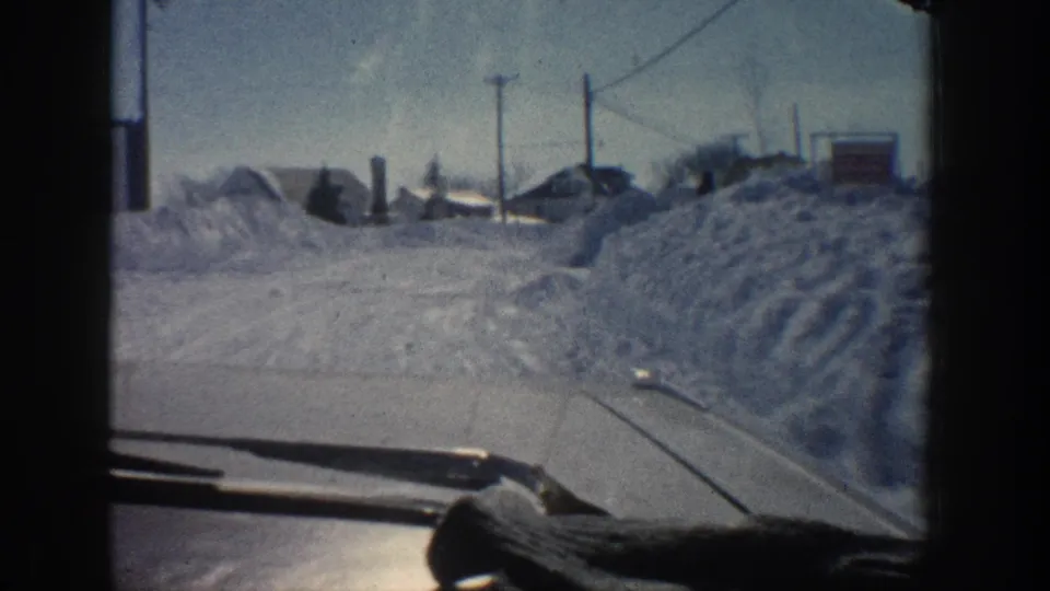 1961 Aspen home-movie view through a car windshield along a snow-covered road bordered by high snowbanks, houses, and utility poles.