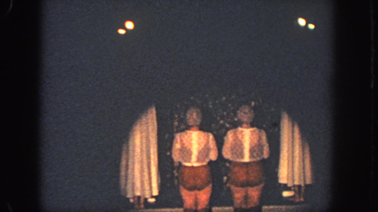 In 1951 Atlanta home-movie footage, two performers in white tops and brown shorts stand on a dark stage between pale draped curtains.