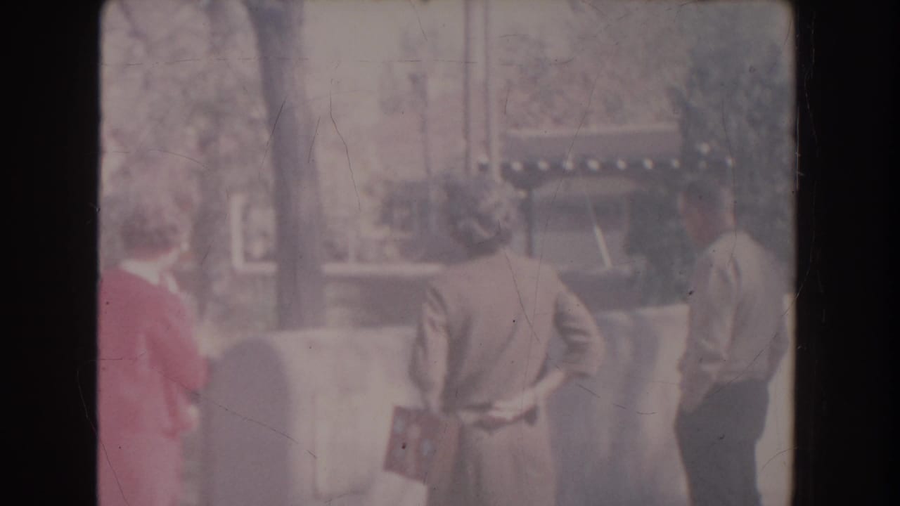 Three adults stand outdoors near a low wall, facing away toward an unseen subject.