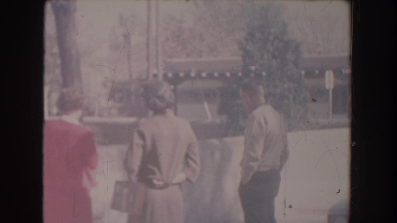 Adults stand in sunlight beside a wall, with trees and a building behind them.