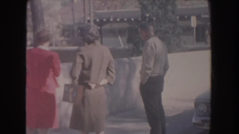 A 1951 home-movie frame shows a woman with a small bag between two adults, all standing near a wall in bright outdoor light.