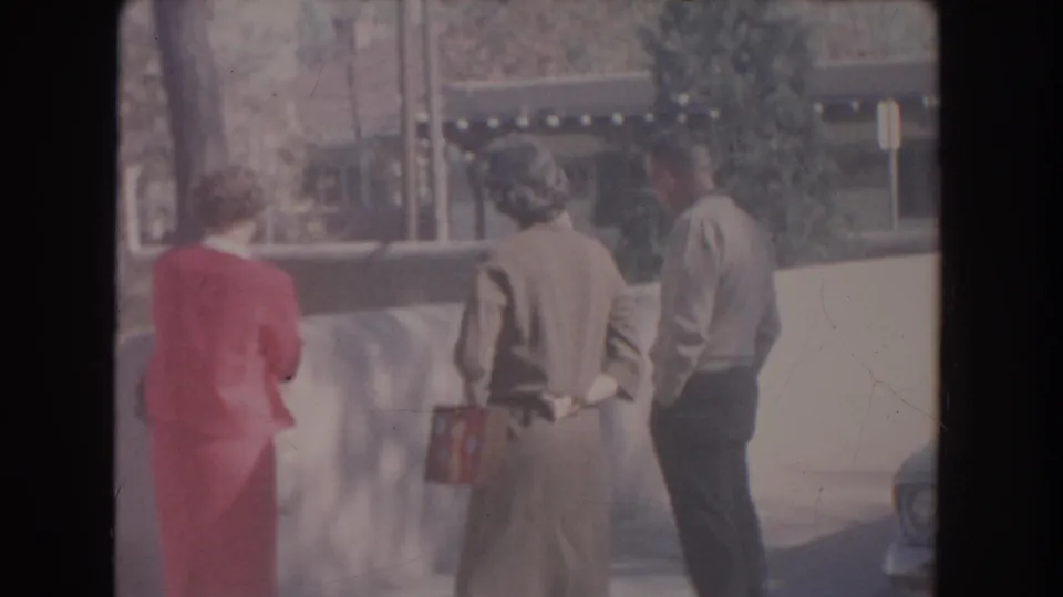 Final frame of the short 1951 New Mexico scene, with three adults near a wall and a partial parked car at the frame edge.