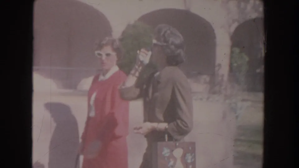 The women stand close together near an arched exterior, with the woman in red wearing white sunglasses and the woman in brown holding a patterned purse.