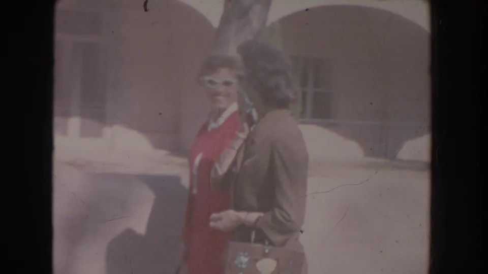 A woman in a red dress and white sunglasses stands beside a woman in a brown suit carrying a patterned purse near a sunlit arched exterior.