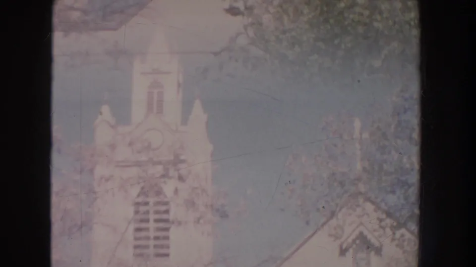 1951 New Mexico home-movie frame showing a white church steeple and tower through soft focus and tree cover.