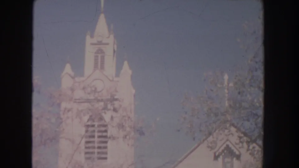 1951 New Mexico home-movie frame with a white church steeple, upper facade, and nearby trees under bright daylight.
