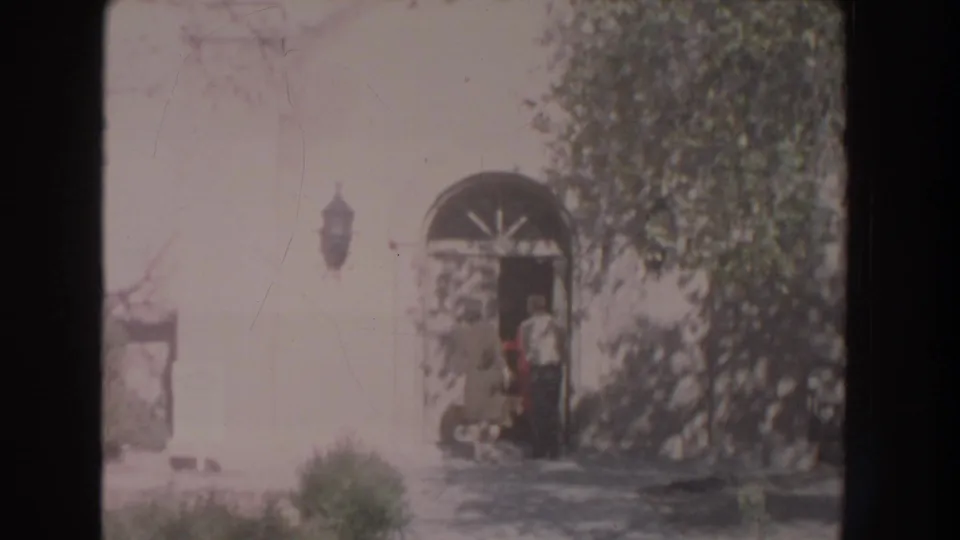 1951 New Mexico home-movie frame showing a person near an arched doorway beside a sunlit white building and leafy trees.