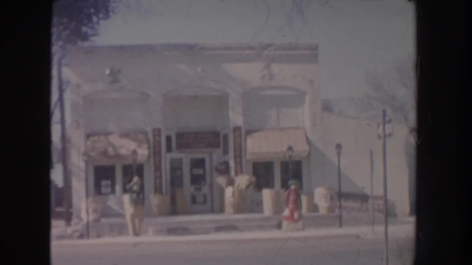 1951 New Mexico home-movie frame showing a small-town storefront row with striped awnings, sidewalk, streetlamp, and distant people.