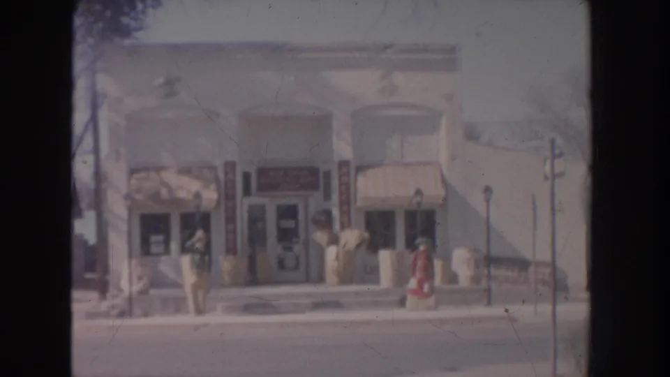 1951 New Mexico home-movie frame showing a storefront across the street with striped awnings, pedestrians, bare trees, and a lamppost.