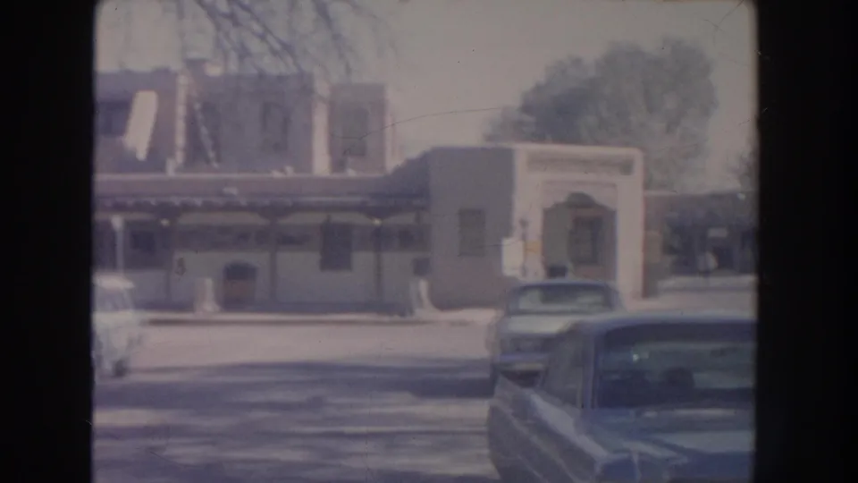 1951 New Mexico home-movie frame showing parked cars along a street facing a commercial building with awnings and a recessed doorway.