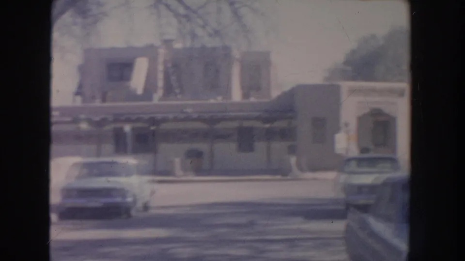 1951 New Mexico street frame with two parked cars, a low commercial building, striped awnings, bare trees, and soft 8mm film edges.
