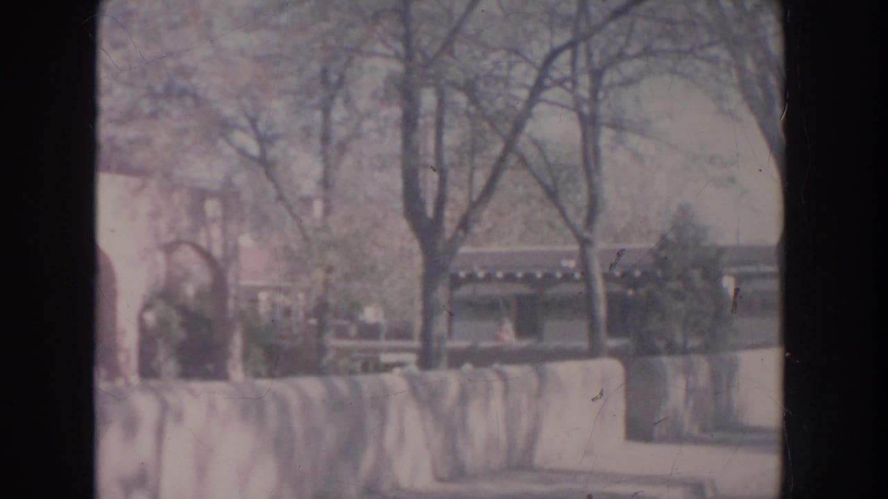 Soft 8mm view of adobe-style arches, a low stone wall, bare trees, and shrubs.