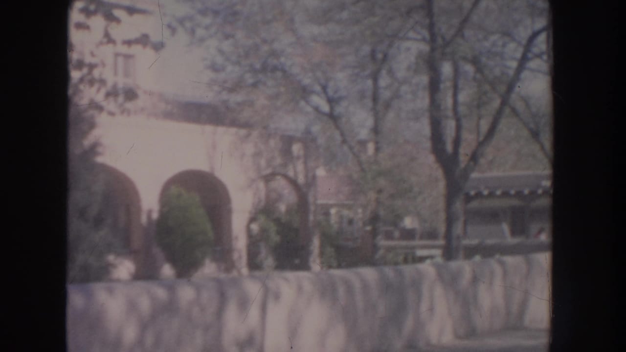 Adobe-style archways, bare trees, shrubs, and a low wall seen in faded 8mm color.
